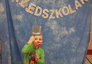Chłopiec uśmiecha się do zdjęcia z papierowym kapeluszem, w tle dekoracja dzień przedszkolaka