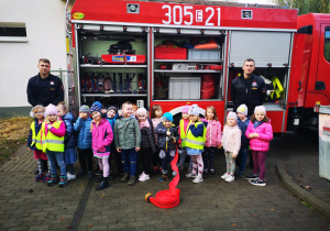 Grupa dzieci pozuje do zdjęcia ze strażakami w tle wóz strażacki