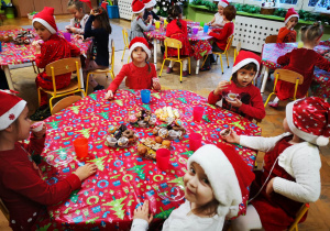 Dzieci w strojach z akcentem świątecznym siedzą przy stoliku, na stoliku kolorowy obrus i słodycze