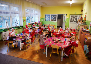 Dzieci w strojach z akcentem świątecznym siedzą przy stolikach, na stolikach kolorowe obrusy i słodycze