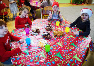 Dzieci w strojach z akcentem świątecznym siedzą przy stoliku, na stoliku kolorowy obrus i słodycze