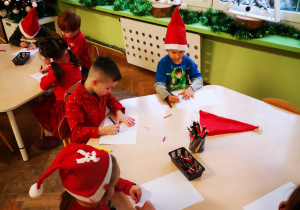Dzieci w mikołajowych czapach rysują przy stolikach