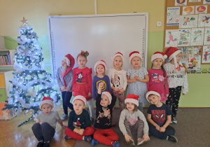 Grupa dzieci w czapkach św. Mikołaja pozuje do zdjęcia przy choince