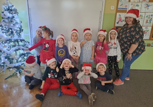 Grupa dzieci z nauczycielką w czapkach św. Mikołaja pozuje do zdjęcia przy choince