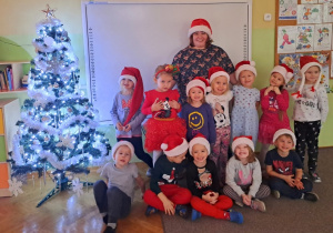 Grupa dzieci z nauczycielką w czapkach św. Mikołaja pozuje do zdjęcia przy choince