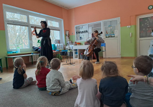 Dzieci siedzą na dywanie i słuchają koncertu, muzyk gra na instrumencie