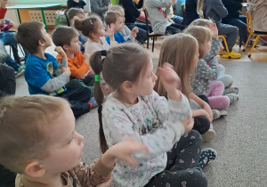 Dzieci siedzą na dywanie i naśladują ruchy pokazywane przez osobę prowadzącą koncert