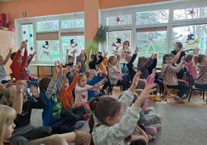Dzieci siedzą na dywanie i naśladują ruchy pokazywane przez osobę prowadzącą koncert