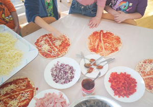 Dzieci siedzą przy stolikach i układają składniki na cieście posmarowanym sosem