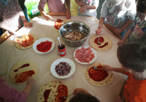 Dzieci stoją przy stoliku i smarują sosem pomidorowym ciasto na pizze