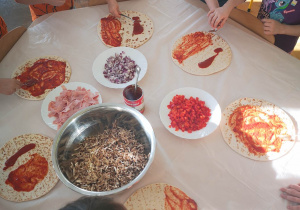 Dzieci stoją przy stoliku i smarują sosem pomidorowym ciasto na pizze