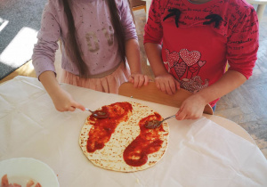 Dziewczynki stoją przy stoliku i smarują sosem pomidorowym ciasto na pizze