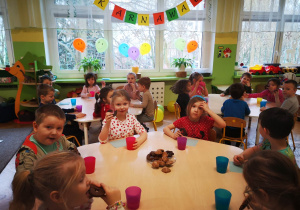 Dzieci siedzą przy stolikach i jedzą słodkie ciasteczka, na stolikach stoją kolorowe kubki