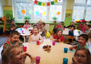 Dzieci siedzą przy stolikach i jedzą słodkie ciasteczka, na stolikach stoją kolorowe kubki