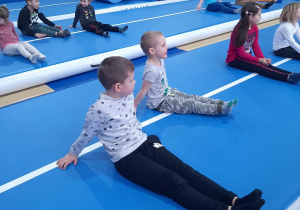 Dzieci siedzą w siadzie prostym na materacach w sali gimnastycznej