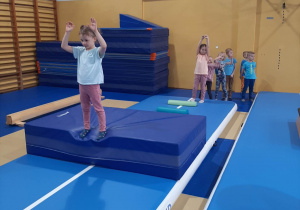 Dzieci wykonują ćwiczenia gimnastyczne na materacach, w tle drabinki, kosz do koszykówki