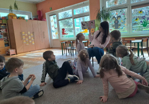 Dzieci siedzą na dywanie, kobieta pokazuje dzieciom ilustracje z książki