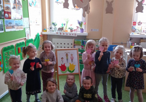 Grupa dzieci pozuje do zdjęcia, dziewczynki trzymają papierowe serca w tle tablica z ilustracjami i dekoracje wielkanocne