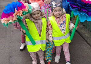 Dzieci w parach stoją na chodniku. Dziewczynki trzymają kolorowe kwiaty z bibuły.