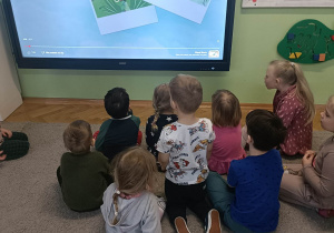 Dzieci siedzą na dywanie i oglądają film edukacyjny na monitorze