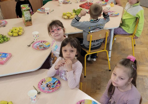 Dzieci siedzą przy stolikach, na stolikach stoją owoce