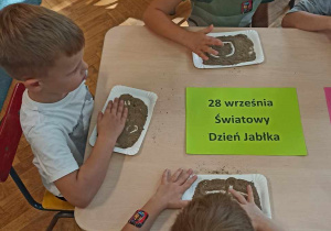 Dzieci siedzą przy stoliku i rysują na piasku kształt jabłka