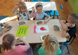 Dzieci siedzą przy stoliku i rysują na piasku kształt jabłka