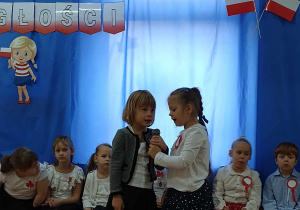 Dziewczynki mówią przez mikrofon wiersze podczas obchodów Święta Niepodległości. W tle dekoracja patriotyczna