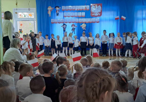 Dzieci ubrane na galowo stoją i śpiewają piosenkę o tematyce patriotycznej, przed nimi widownia macha flagami w tle dekoracja tematyczna