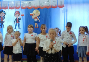 Dziewczynka mówiąprzez mikrofon wiersz podczas obchodów Święta Niepodległości inne dzieci stoją za nią. W tle dekoracja patriotyczna