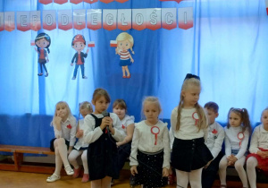 Dzieci mówią przez mikrofon wiersze podczas obchodów Święta Niepodległości. W tle dekoracja patriotyczna