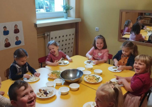 Dzieci siedzą przy stole i zjadają sałatkę owocową.