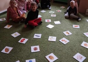 Dzieci siedzą na dywanie i oglądają ilustracje produktów znajdujących sie na dywanie.