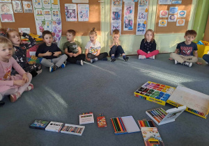 Dzieci siedzą na dywanie i słuchają opowiadania. Na środku leżą różne rodzaje kredek w pudełkach