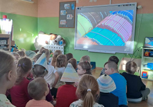 Dzieci siedzą na podłodze i oglądają na tablicy multimedialnej film z fabryki kredek