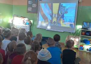 Dzieci siedzą na podłodze i oglądają na tablicy multimedialnej film z fabryki kredek