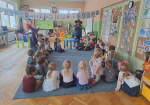 Dzieci siedzą na dywanie, nauczycielka w kapeluszu opowiada o tradycjach andrzejkowych
