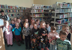 Dzieci w bibliotece stoją miedzy półkami z książkami i słuchają o pracy bibliotekarza