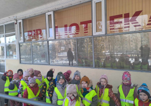 Grupa dzieci w kamizelkach odblaskowych pozuje do zdjecia przed wejściem do biblioteki