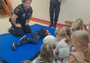 Strażniczka Miejska prezentuje i objaśnia sposób układania poszkodowanego w pozycji bezpiecznej dzieci patrzą.