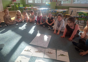 Dzieci siedzą na dywanie i słuchają informacji o dinozaurach
