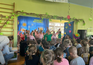 Grupa dzieci stoi na scenie przed widownią i śpiewa piosenkę. W tle dekoracja wiosenna
