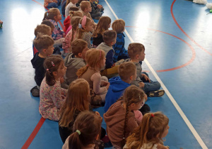 Dzieci siedzą na podłodze i oglądają przedstawiany przez uczniów