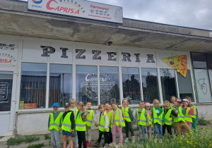 Grupa dzieci w kamizelkach odblaskowych pozuje do zdjęcia pod budynkiem pizzerii