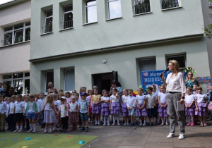 Dzieci ustawione przed budynkiem oczkują na występ, nauczycielka przemawia przez mikrofon