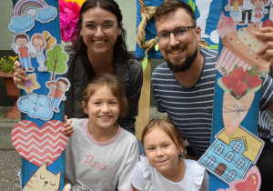 Rodzice z dziećmi pozują do zdjęcia w ramce z napisem Dzień Rodziny i obrazkami tematycznymi