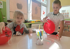 Dzieci siedzą przy stoliku i ozdabiają kolorowymi mazakami czerwone balony.