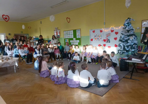 Dzieci siedzą w kole. Goście siedzą na widowni.